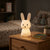 Little Bunny Silicone Night Light glowing on a bedside table with books and stuffed animals in a cozy bedroom.