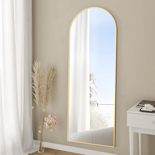 Arched Full Length Floor Mirror with Gold Frame leaning against a beige wall, reflecting natural light and creating a spacious feel.