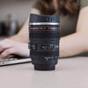 Camera Lens Travel Mug with Lid on a table next to a laptop, resembling a real camera lens.