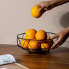 Geometric Iron Fruit Storage Basket with fresh oranges being placed inside on a wooden table.
