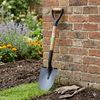 Heavy-Duty Wooden Handle Garden Shovel leaning against a brick wall in a garden setting with flowers.