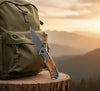 3D Pattern Blade Olive Wood Handle Pocket Knife next to a backpack on a log in a scenic outdoor setting.