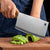 Heavy Duty Stainless Steel Bone Chopping Butcher Cleaver slicing through green vegetables on a wooden cutting board.