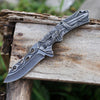 3D Embossed Dragon Folding Survival Knife resting on wooden surface, showcasing intricate handle design and sharp blade.