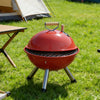 Portable Stainless Steel Charcoal BBQ Grill in bright red color set up on grassy area with camping equipment nearby.