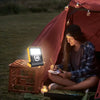 A woman sitting by a tent using the Solar LED Work Light Rechargeable Waterproof while writing outside at night.