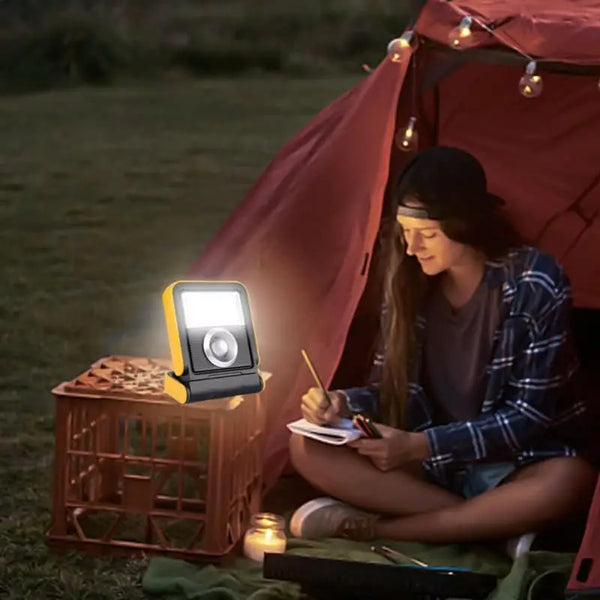 A woman sitting by a tent using the Solar LED Work Light Rechargeable Waterproof while writing outside at night.