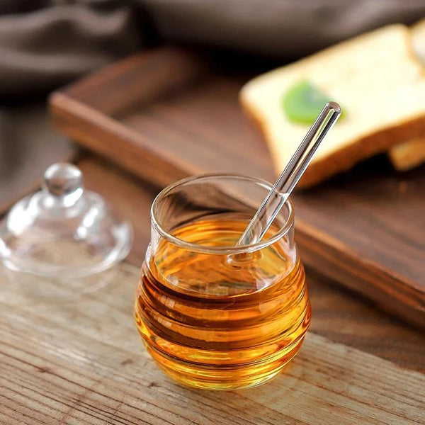 Hand-Blown Bee-Shaped Borosilicate Glass Honey Jar with honey and dipper beside a slice of bread.