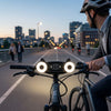 3-in-1 Smart Outdoor Floodlight Security Camera Unit: Person riding a bicycle at dusk with bright front lights on the handlebar in a city setting.