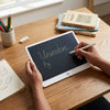 User writing on an LCD Writing Tablet Drawing Board with a stylus on a wooden desk.