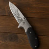 Engraved Wilderness Tactical Folding Knife featuring a detailed blade design and sturdy handle on a wooden surface.