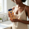 Woman holding Cocoa Butter Glow Body Oil Gel bottle in a bright kitchen.