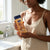 Woman holding Cocoa Butter Glow Body Oil Gel bottle in a bright kitchen.