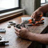 Person using Bent Nose Pliers to grip and manipulate mechanical parts on a wooden workbench.
