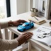 Person playing with a blue Die-Cast Racing Car Toy on a wooden table next to a magazine and decorative items.