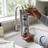 Person holding a Faucet Sprayer package beside a kitchen sink with a 360 Degree Rotating Faucet Anti-Splash Filter.