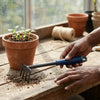 A person using a Five Teeth Garden Harrow Rake to cultivate soil around a potted plant in a greenhouse.