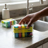 Person reaching for a pack of colorful Dual-Action Kitchen Cleaning Sponges on a kitchen countertop.