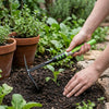 Using a Multifunction Telescopic Garden Hoe and Fork to tend to plants in a garden bed with potted herbs.