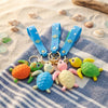 Colorful 3D Rubber Sea Turtle Keychain collection featuring vibrant turtle designs and blue straps on a sandy background.