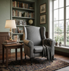 Serena Grey Fabric Wingback Chair in a cozy living room setting next to a wooden table and bookshelf.