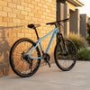 Mountain Bike with Suspension Fork & Triple Chainset leaning against a brick wall in a serene outdoor setting.