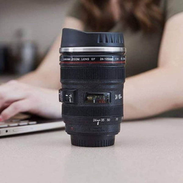 Camera Lens Travel Mug with Lid on a table next to a laptop, resembling a real camera lens.
