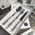 Stainless Steel Cutlery Set displayed in a box featuring a spoon, fork, and knife with stylish black and white handles.