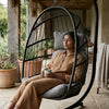 Woman relaxing in the Steel Frame Hanging Egg Chair with a mug, surrounded by plants in a cozy outdoor setting.