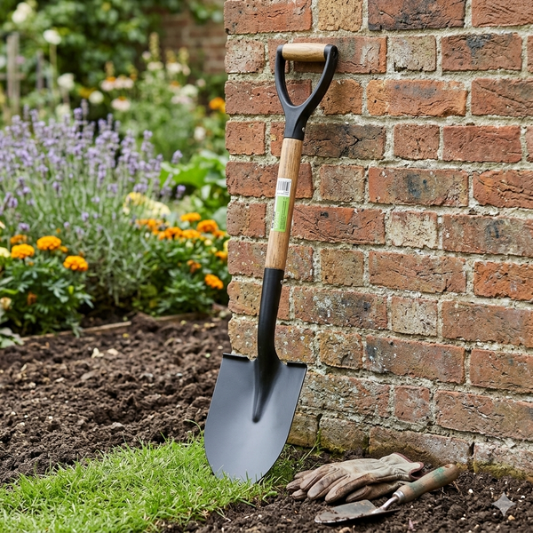 Heavy-Duty Wooden Handle Garden Shovel leaning against a brick wall in a garden setting with flowers.