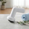 Handheld Body Massager resting on a countertop next to a rolled blue towel and green plant in a bright room.