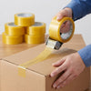 Person applying Secure Seal Clear Packing Tape Roll on a cardboard box with multiple tape rolls in the background.