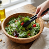 A hand using Multifunction Stainless Steel Locking Food Tongs to serve a fresh salad in a wooden bowl.