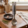 Stainless Steel Speckled Handle Serving Ladle on a wooden counter with cooking utensils and herbs.