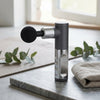 Portable Mini Massage Gun on a countertop next to a towel and greenery, showcasing its compact design.