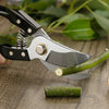 Steel Garden Bypass Pruner cutting a green branch on a wooden surface, demonstrating its heavy-duty design.