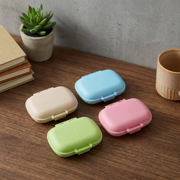 Four colorful Weekly Pill Organizer Cases in pastel colors on a wooden table next to a plant and stack of notebooks.