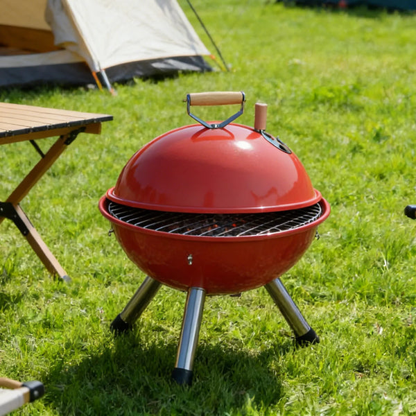 Portable Stainless Steel Charcoal BBQ Grill in bright red color set up on grassy area with camping equipment nearby.