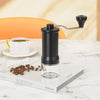 Manual Ceramic Coffee Grinder on a table with coffee beans and a cup of coffee in the background.