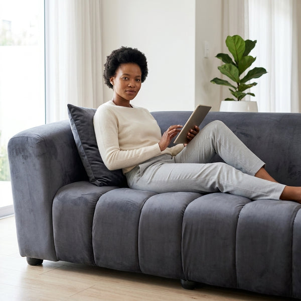 Modern Grey Upholstered 2-Seater Sofa with a woman relaxing and using a tablet in a bright living space.