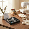 11-Piece Sewing Machine Presser Feet Kit displayed on a wooden table with sewing supplies and a sewing machine in the background.