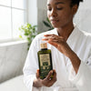 Woman in a robe holding a bottle of Olive Oil Infused with Argan Oil & Rose Petals for hair health.