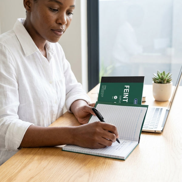 A6 Feint Ruled Duplicate Book Pack being used by a woman writing notes at a desk with a laptop.