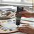 A hand holding a tube of Acrylic Paint 75ml - Olive Green, used for mixing colors on a wooden palette.