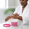 Woman applying Tropical Musk Skin Treatment 1L moisturizer from a tub for hydrated skin.