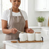 Woman in apron using Ceramic Seasoning Jar Set with Wooden Tray to measure spices in a modern kitchen.
