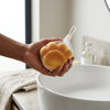 Soft Slow Rising PU Butter Stick Squishy Toy: Person holding a mesh bag of sponge balls near a wash basin in a bright kitchen.