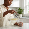 Woman pouring from a High Borosilicate Glass Cold Water Kettle into a glass with lemon slices.