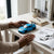 Person playing with a blue Die-Cast Racing Car Toy on a wooden table next to a magazine and decorative items.