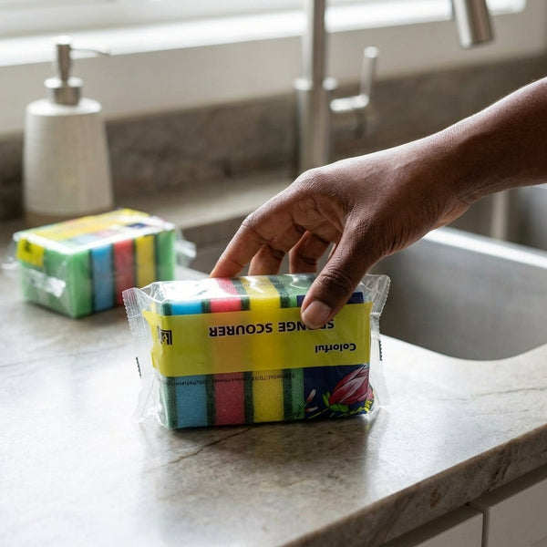 Person reaching for a pack of colorful Dual-Action Kitchen Cleaning Sponges on a kitchen countertop.
